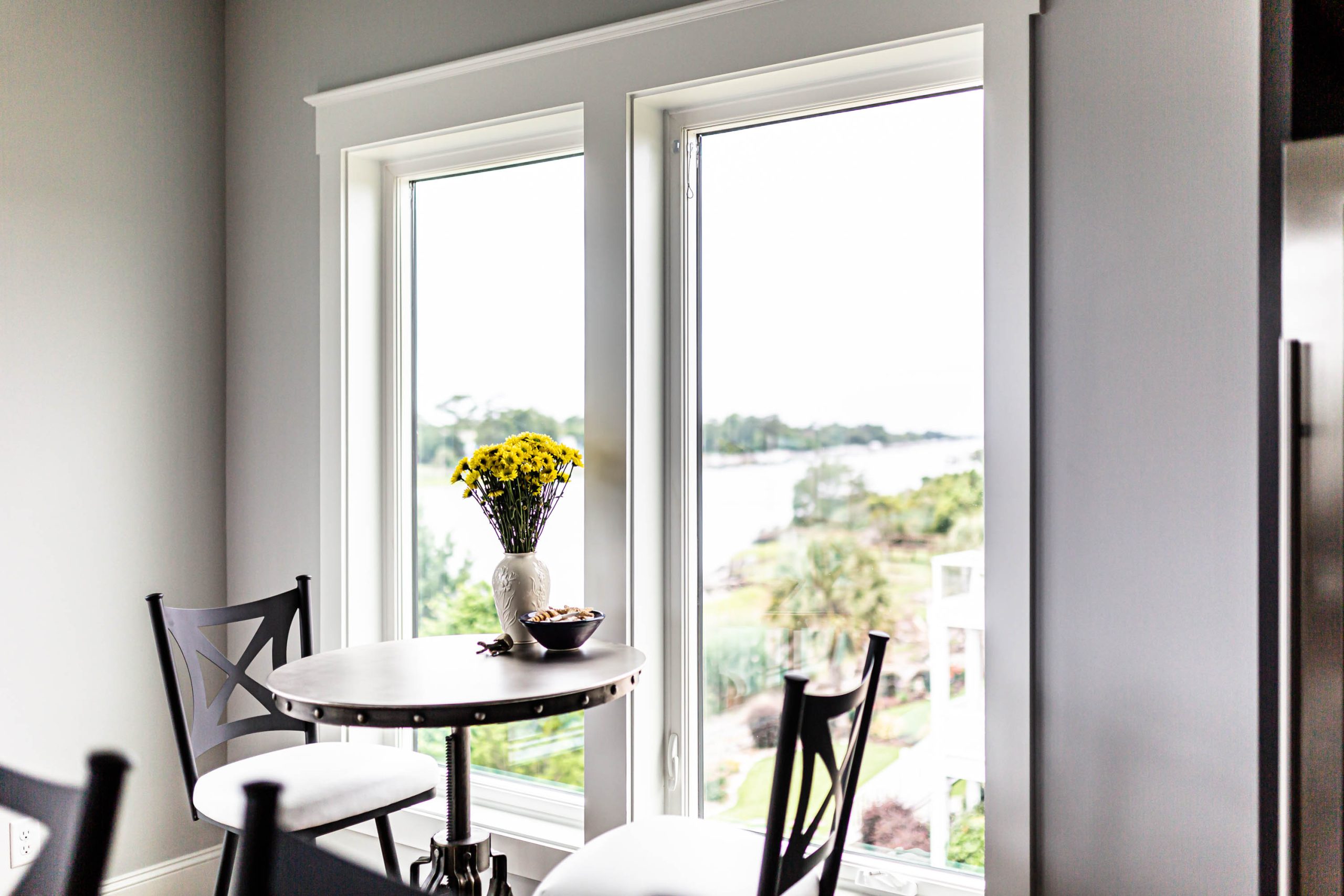 Mulled picture windows sitting in front of a small table with a bouquet of yellow flowers, along with a cup. Two chairs sit at the table.
